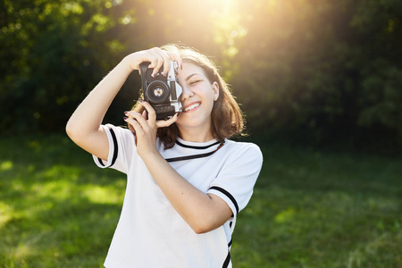 在绿色公园或草地上 , 穿着白色上衣拍照的微笑可的肖像。年轻女性摄影师与老式相机拍摄日落
