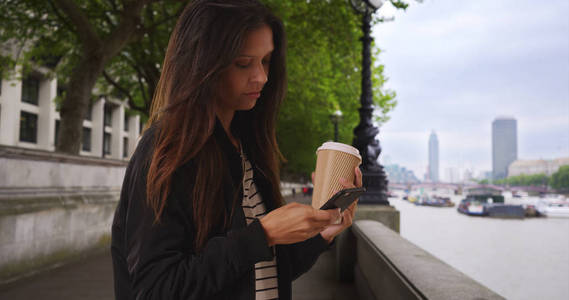 伦敦的 somber 女性拿着她的咖啡在泰晤士河边用手机发短信