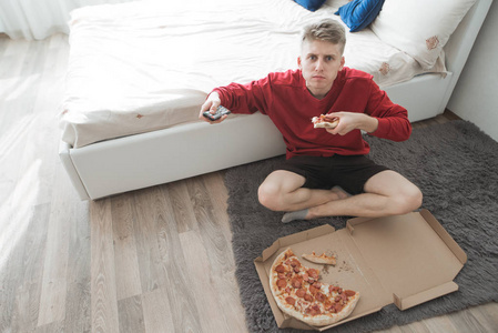 一个严肃的年轻人坐在家里的房间里, 地板上拿着一盒比萨饼, 看着相机, 吃比萨饼。青少年吃比萨饼从盒子里看着相机