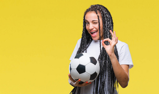 年轻的编织头发非洲裔美国女孩拿着足球在孤立的背景做确定的标志与手指, 优秀的象征