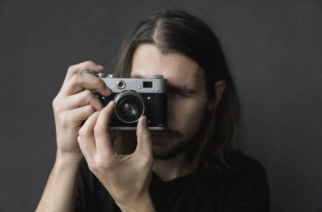 英俊的年轻胡须男子, 长头发, 穿着黑色衬衫, 在黑色背景上拿着老式老式电影相机, 在相机取景器中寻找