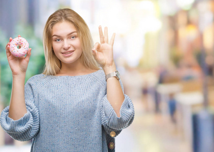 年轻的白种女人吃甜甜甜圈在孤立的背景做 ok 符号与手指, 优秀的符号