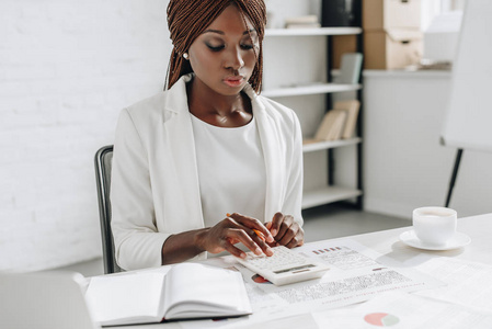 专注非洲裔美国成年女商人在白色正式服装在办公桌工作, 并使用计算器