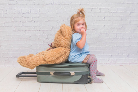 女孩, 在白色砖墙背景下的孩子坐在一个手提箱, 他的背部转向玩具熊