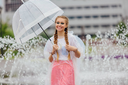 年轻漂亮的女孩与两个辫子在黄色靴子和透明的雨伞站在喷泉附近。城市的雨天