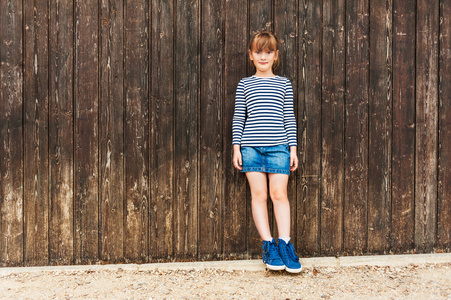一个可爱的小女孩，穿着连衣裙 牛仔裙 美丽的蓝色运动鞋，棕色木墙站的室外肖像