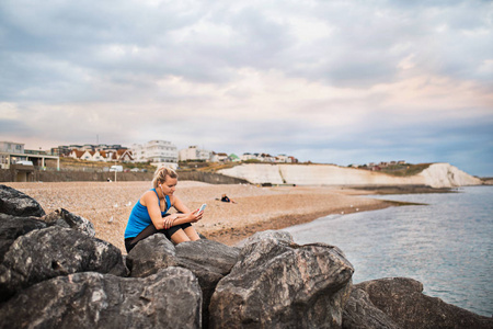 年轻运动妇女赛跑者与耳机和智能手机坐在外面在海滩在自然, 听音乐和休息