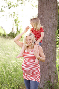 孕妇抱着女儿在草地上的树附近的肩膀上