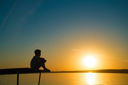男孩看着日落小男孩坐在一座桥上, 看着河边的日落.