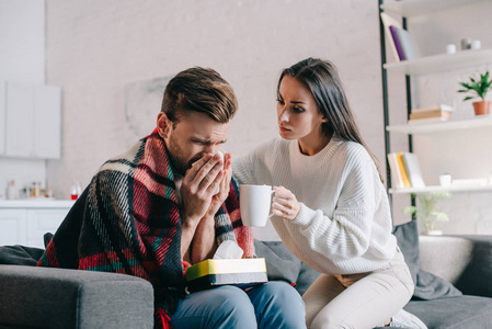 美丽的年轻女子拿着一杯温暖的饮料生病的男朋友, 而他打喷嚏