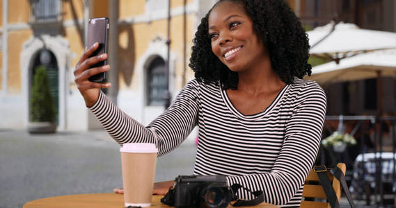 微笑的黑人妇女坐在罗马的户外咖啡馆拿着手机自拍