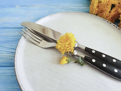 在蓝色木质背景上用黄色菊花制成的叉子盘子小刀