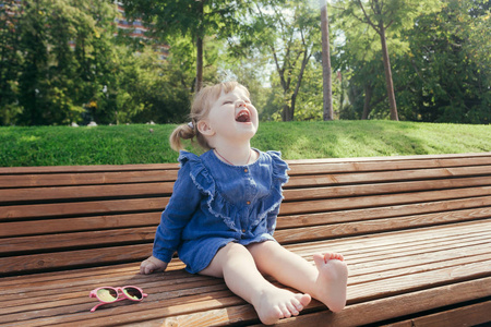 小女孩坐在长椅上, 在一个温暖的阳光明媚的日子在城市公园里欢笑