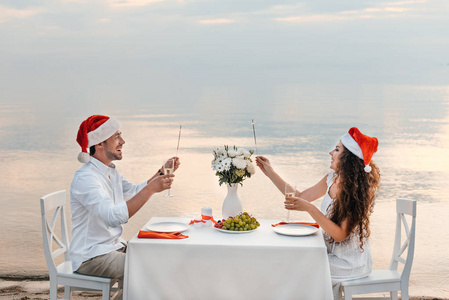 快乐夫妇在圣诞老人的帽子庆祝圣诞节与香槟眼镜和烟花在海滩上