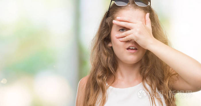 年轻的金发女郎戴着太阳镜偷看着脸和眼睛用手看着通过手指尴尬的表情