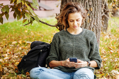 美丽的年轻微笑的女孩, 波浪般的头发坐在草地上, 秋天的叶子与智能手机在手