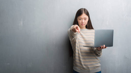 年轻的中国妇女在灰色墙壁使用计算机手提电脑指向照相机和对您, 手标志, 正面和自信手势从前面