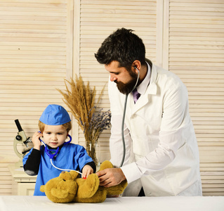 兽医和小助手检查泰迪熊。父亲和孩子细心面对扮演医生。保健和儿童概念。男子和男孩在木质背景上按住听诊器