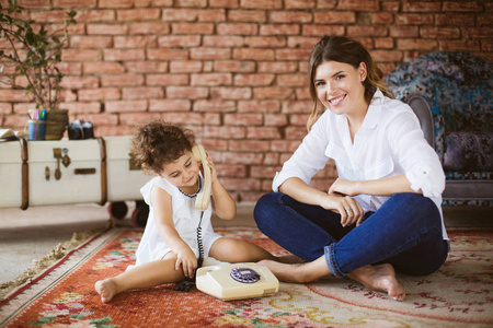 美丽的微笑的母亲高兴地看着相机, 而小可爱的女儿朦胧地玩复古电话附近的地毯在家