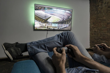 两个男人在玩电子游戏。手持控制台控制器。电视上的足球或足球比赛。宽屏电视挂在墙上