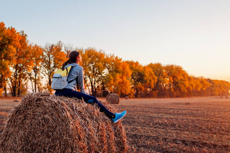 年轻的旅行者在秋天的田野休息日落。妇女欣赏视图坐在干草堆。运动娱乐概念