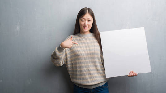 年轻的中国女人在灰色的墙壁上拿着横幅与惊喜的脸指向自己