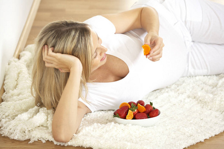 愉快的怀孕妇女与碗草莓和桔子躺在地毯在家里