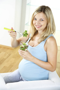 愉快的怀孕妇女坐在扶手椅和吃沙拉从玻璃在家