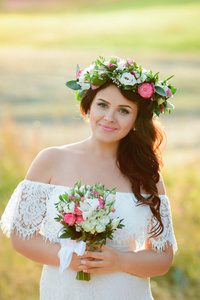 美丽的黑发女孩与花束和花环的花朵。在白色蕾丝连衣裙长卷发的年轻妇女。夏季女性肖像日落