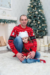 愉快的爸爸和女儿在红色毛衣坐在地毯附近的壁炉装饰的圣诞前夜