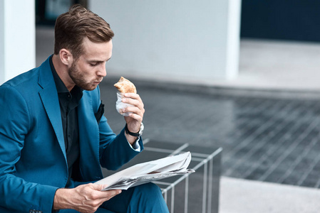 获取信息。忙碌的年轻英俊商人坐在办公楼附近吃羊角面包和看报纸