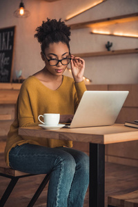 美丽的美国黑人女孩在休闲服装和眼镜正在与笔记本电脑在咖啡馆