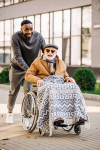快乐的高级残疾人在轮椅上与格子和非洲裔美国男子骑在街道