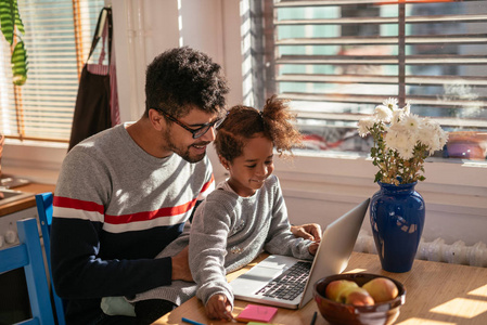 一张美国父亲和女儿在室内玩电脑的照片。她坐在他的大腿上。他们都在微笑