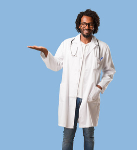英俊的非洲裔美国医生手持东西, 显示一个产品, 微笑和欢快, 提供一个假想的对象