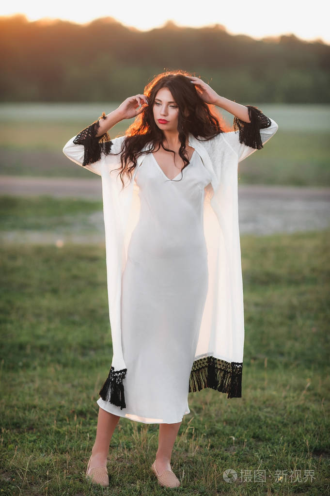 美丽的卷曲的女孩在松散的衣服上的自然日落背景