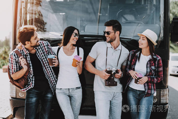 年轻人在旅游巴士前喝咖啡.一群带着背包站在一起聊天的微笑的朋友.