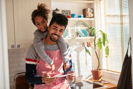 父亲和女儿粘接在厨房里一边读食谱在平板电脑上。那个男人戴着围裙，女孩从后面拥抱他。他们都在笑