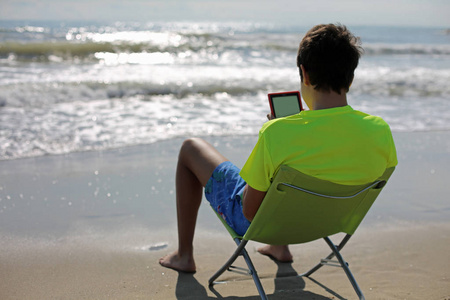 少年读一本数字书坐在一张折叠的椅子在海前面