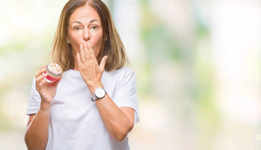 中年西班牙妇女吃纸杯蛋糕在孤立的背景覆盖嘴与手震惊的耻辱, 表达恐惧, 害怕在沉默中, 秘密的概念