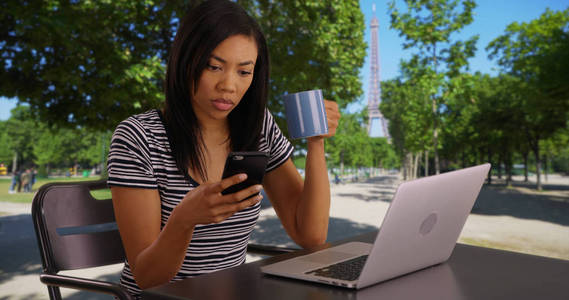 在法国巴黎公园里, 黑人妇女一边喝咖啡, 一边用智能手机发短信
