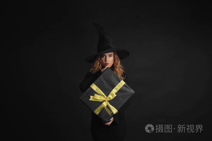 黑色星期五。万圣节女巫与黑色的帽子在黑色背景拿着礼物盒与朱斯在她的手中。惊喜和礼品在售