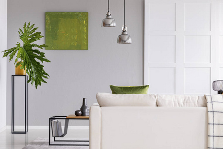 白色沙发上方的灯在简单的客厅内部, 桌子上有绿色的绘画和植物。真实照片