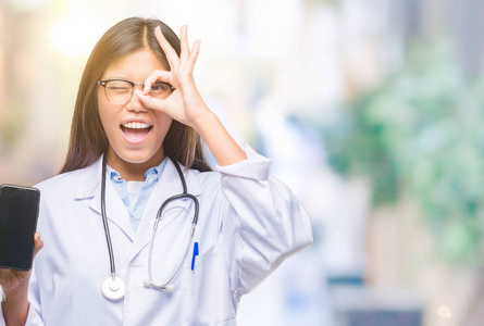 年轻的亚洲医生妇女持有智能手机在孤立的背景下与快乐的脸微笑做 ok 标志与手在眼睛看通过手指