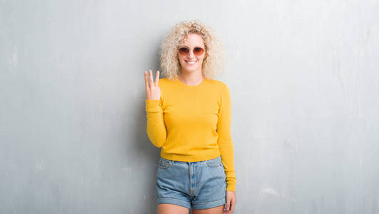 年轻的金发女人与卷曲的头发在垃圾灰色背景显示, 并指出手指三, 而微笑着自信和快乐