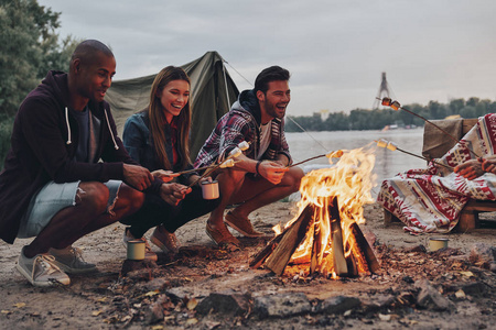 一群快乐的人在休闲穿着烤棉花糖在篝火边休息在湖附近