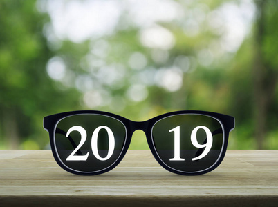 2019白色文本与黑眼睛眼镜在木桌上模糊绿色树在公园, 商业愿景, 新年快乐2019概念
