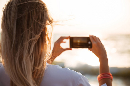 在海滩上用手机拍摄日落照片的妇女的后视图