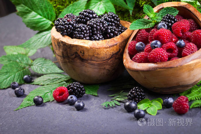 两个木碗的食物组成蓝莓, 黑莓和树莓与绿叶的灰色背景