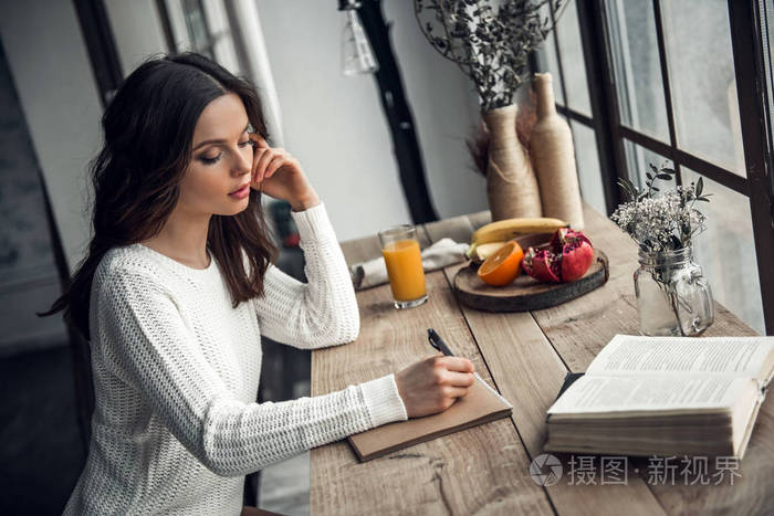 美丽的年轻女孩在休闲服装是写在记事本上, 而坐在木桌在家里